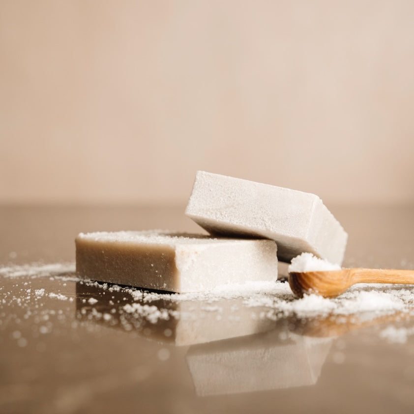 Two sea salt soap bars on a reflective surface with a wooden spoon of sea salt.