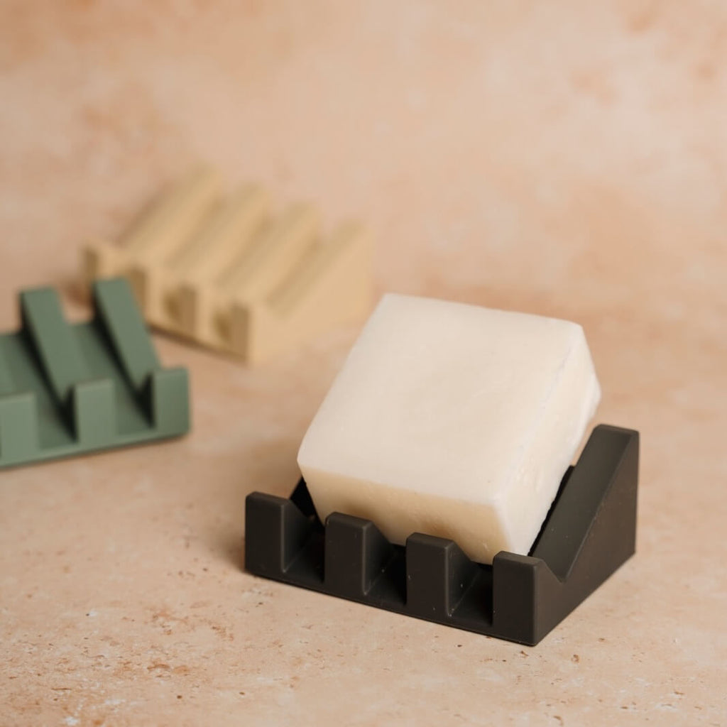 Bar of soap in a black soap dish on a beige surface with a blurred brush in the background.