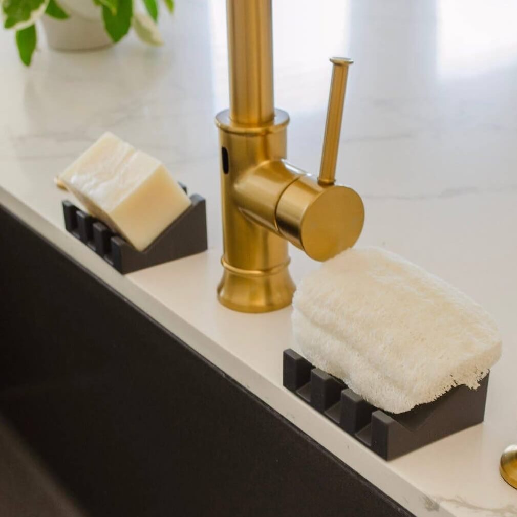 Gold faucet with black soap dish and sponge holder on a bathroom counter.